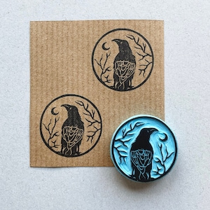 May include: Three rubber stamps featuring a black crow design within a circle. The crow is facing left and has a crescent moon behind it. The stamps are displayed on a brown paper background.