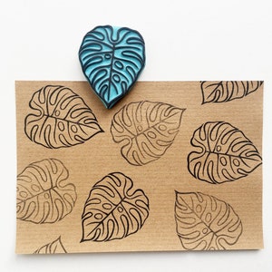 May include: A sheet of brown paper with a repeating pattern of black monstera leaves. A blue rubber stamp of a monstera leaf is on top of the paper.