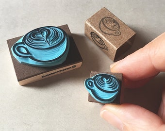 May include: A selection of coffee-themed stamps. The stamps depict a latte art design in a light blue hue. One stamp is mounted on a brown block, and another is held by a hand. A small brown box with a coffee cup design is also present.