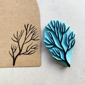 Puede incluir: Un sello de goma azul con un dise&ntilde;o de rama de &aacute;rbol negro junto a un papel marr&oacute;n con el mismo dise&ntilde;o estampado con tinta negra. El sello tiene forma de hoja y est&aacute; sobre una superficie blanca.