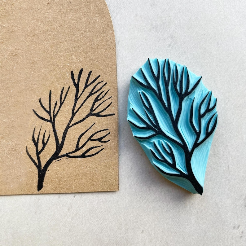 Puede incluir: Un sello de goma azul con un dise&ntilde;o de rama de &aacute;rbol negro junto a un papel marr&oacute;n con el mismo dise&ntilde;o estampado con tinta negra. El sello tiene forma de hoja y est&aacute; sobre una superficie blanca.