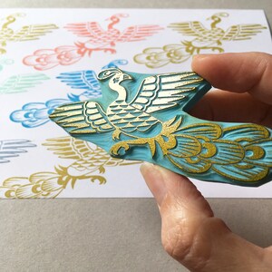 May include: A blue rubber stamp with a gold design of a stylized bird with a long tail. The bird is in a flying pose with its wings spread out.