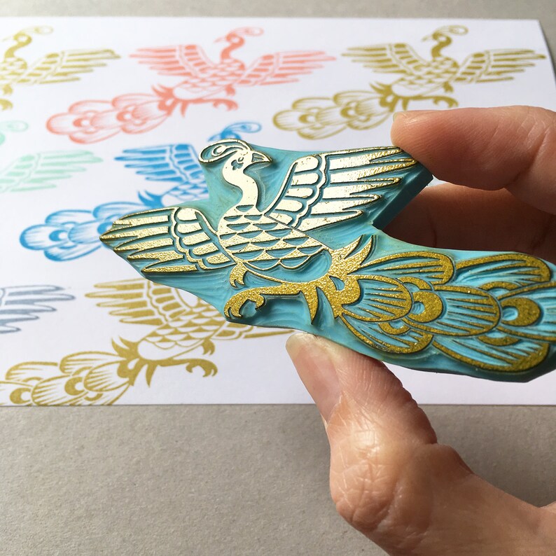 May include: A blue rubber stamp with a gold design of a stylized bird with a long tail. The bird is in a flying pose with its wings spread out.
