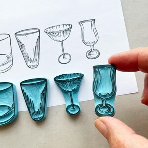 May include: Teal rubber stamps depicting different glassware designs, such as a cocktail glass and a champagne flute, with black outlines. The stamps are held near a paper with matching line drawings of the glassware.