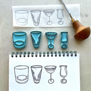 May include: Four blue rubber stamps showing designs of various glasses: a tumbler, a highball, a cocktail glass, and a hurricane glass. A carving tool with a wooden handle and sketches of the glassware are also visible.