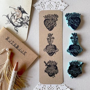 May include: A vertical kraft paper bookmark featuring three black ink stamped heart designs, accompanied by three light blue rubber stamps with matching heart motifs, a notebook with a tassel, a pen, and a card with a cherub illustration.