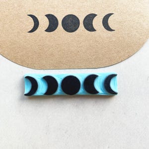 Puede incluir: Un sello de goma azul con un dise&ntilde;o de fases lunares en negro. El sello muestra las fases de la luna en una fila. La imagen estampada tambi&eacute;n se muestra en un trozo de papel marr&oacute;n.