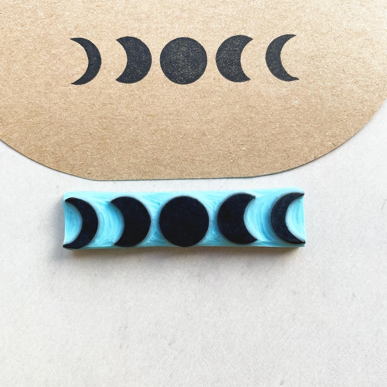 Puede incluir: Un sello de goma azul con un dise&ntilde;o de fases lunares en negro. El sello muestra las fases de la luna en una fila. La imagen estampada tambi&eacute;n se muestra en un trozo de papel marr&oacute;n.