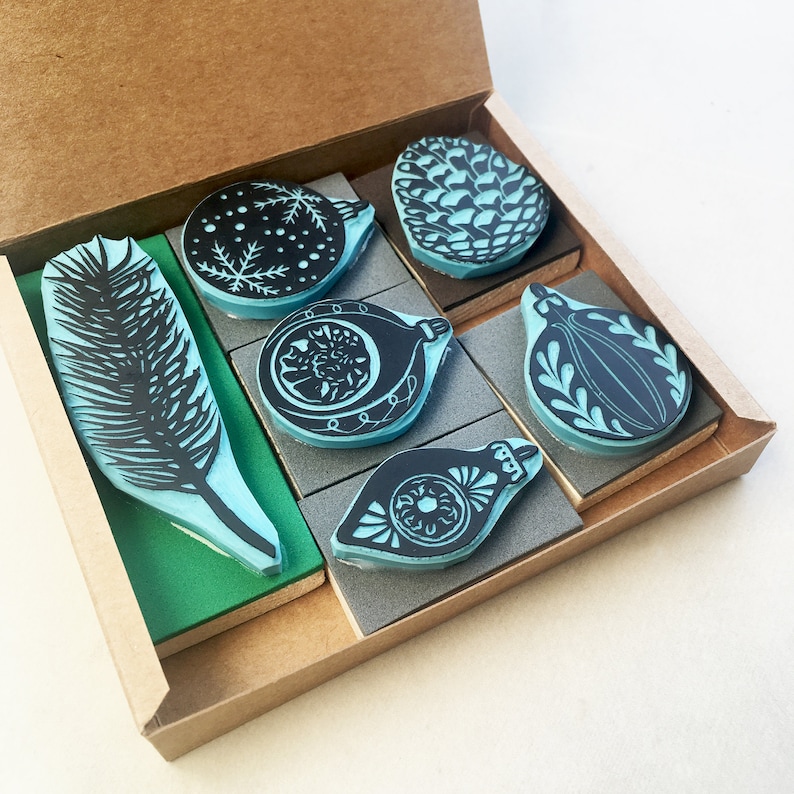 May include: A set of six blue and black rubber stamps in a cardboard box. The stamps feature various holiday designs, including a pine cone, a feather, and ornaments.