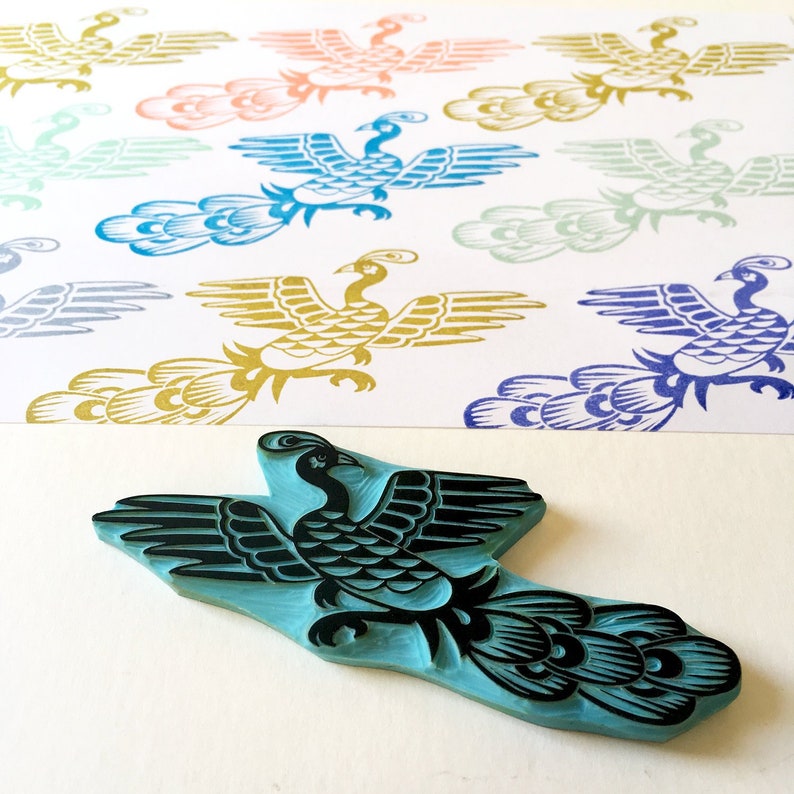 May include: A blue rubber stamp of a stylized peacock with black details. The stamp is on a white surface with several printed images of peacocks in various colors.