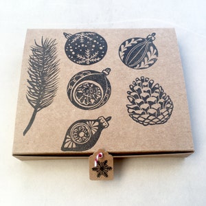 May include: A brown cardboard box with a snowflake tag attached. The box has a black and white design of Christmas ornaments, pine cones, and a pine branch.