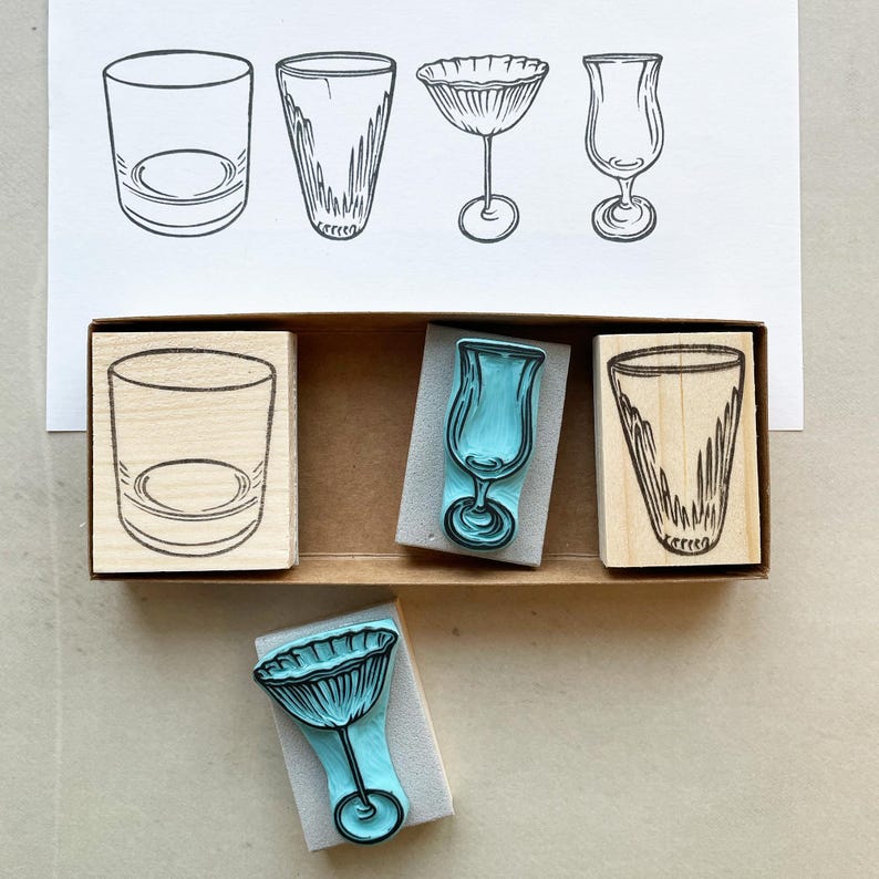 May include: A collection of four drink glass illustrations and four rubber stamps. The stamps are in a wooden box. The stamps depict a rocks glass, a tall glass, a stemmed cocktail glass, and a tulip glass.