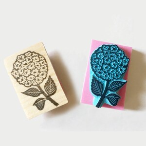 K&ouml;nnte beinhalten: Zwei Blumenstempel. Einer ist ein Holzstempel mit einem schwarzen Hortensien-Design. Der andere ist ein blauer Hortensienstempel auf einem rosa Schaumstoffblock. Die Stempel sind f&uuml;r Bastel- und Kunstprojekte.