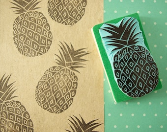 May include: A black and blue pineapple stamp on a green foam pad, next to a paper with repeated pineapple prints. The pineapple stamp has a detailed design, and the prints are in black ink on a tan paper.