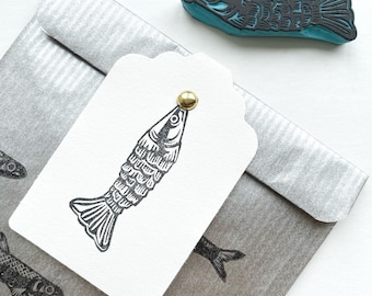 May include: A white gift tag with a black and white illustration of a fish. The tag has a gold brad in the top corner. The tag is on top of a silver envelope with a repeating pattern of black and white fish.