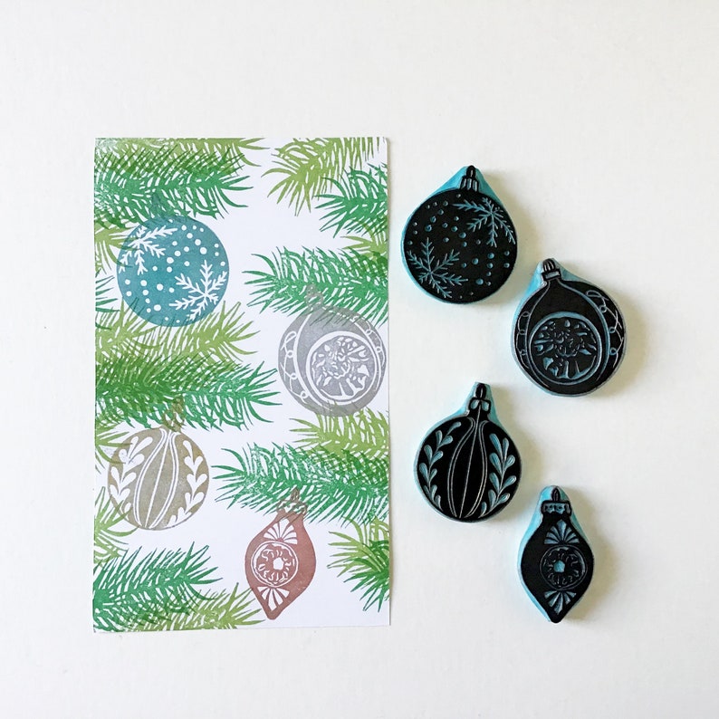May include: A set of six Christmas ornament rubber stamps. The stamps are black and white and feature various designs, including a snowflake, a star, and a bell. The stamps are arranged on a white background with a green pine branch pattern.
