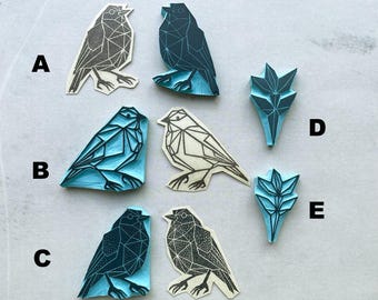 Puede incluir: Una colección de sellos geométricos de pájaros y hojas en varios colores. Los sellos incluyen diseños en gris, verde azulado y azul, con detalles lineales. Los sellos de pájaros están en diferentes poses, y los sellos de hojas tienen una apariencia estilizada.