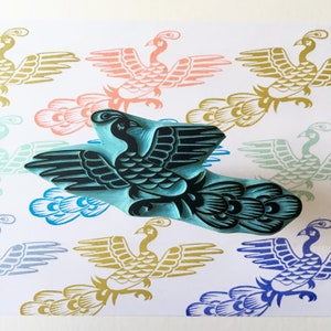 May include: A teal blue wooden block carving of a black and white stylized phoenix bird with a long tail. The carving is on a white background with several printed images of phoenix birds in various colors.
