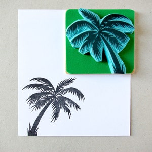 May include: A close-up of a green rubber stamp with a blue palm tree design. The stamp is on a white surface with a black palm tree print.