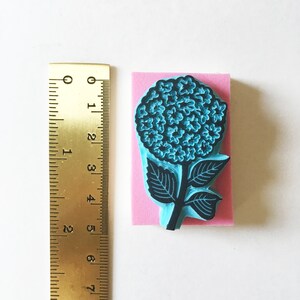 K&ouml;nnte beinhalten: Ein blauer Hortensienbl&uuml;tenstempel mit schwarzen Umrissen, montiert auf einem rosa Schaumstoffblock. Der Stempel ist etwa 7.6 cm hoch. Ein Messinglineal ist zur Ma&szlig;stabsbestimmung neben dem Stempel positioniert.