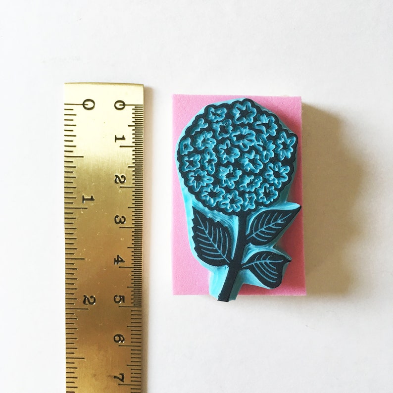 K&ouml;nnte beinhalten: Ein blauer Hortensienbl&uuml;tenstempel mit schwarzen Umrissen, montiert auf einem rosa Schaumstoffblock. Der Stempel ist etwa 7.6 cm hoch. Ein Messinglineal ist zur Ma&szlig;stabsbestimmung neben dem Stempel positioniert.