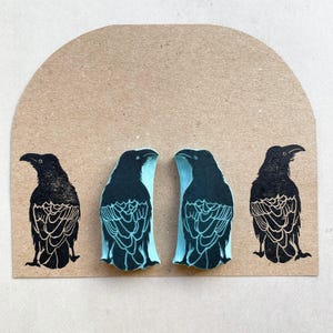 Puede incluir: Cuatro grabados de arte de cuervos negros sobre un fondo marr&oacute;n claro. Dos cuervos se enfrentan con detalles en azul claro, mientras que los otros dos son negros s&oacute;lidos. Los grabados tienen patrones de plumas detallados y est&aacute;n sobre un fondo semicircular.