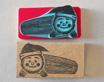 May include: Two woodblock stamps, one blue and one black, depicting a cartoon log with a face and a hat. The blue stamp is on a red background and the black stamp is on a brown background.
