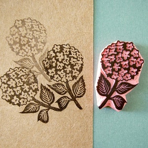 K&ouml;nnte beinhalten: Ein rosa Gummistempel mit einem detaillierten Hortensienbl&uuml;ten-Design. Daneben ein braunes Papier mit einem dunkelbraunen Stempelabdruck der gleichen Blume. Der Abdruck zeigt das Blumenmuster.