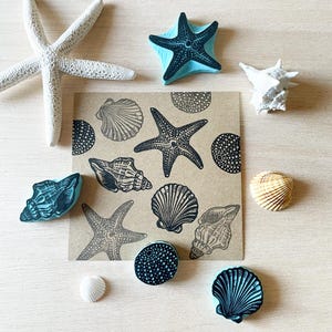 May include: A collection of sea-themed stamps and seashells. The stamps feature starfish and shell designs in teal and black. A white starfish and various seashells are also present, arranged on a light wooden surface.