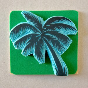 May include: A blue and black palm tree design carved into a green foam block. This is a rubber stamp for crafting and art projects.