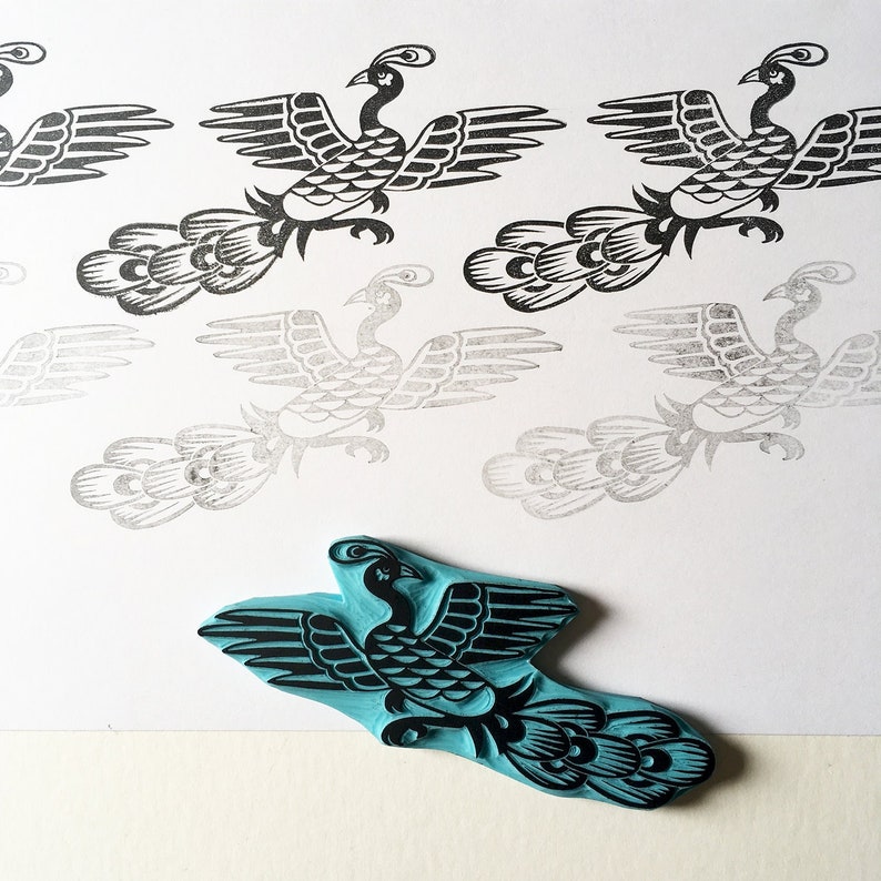 May include: A blue rubber stamp with a black design of a stylized peacock with a long tail. The stamp is on a white surface with several black and gray impressions of the same design.