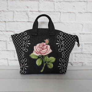 Könnte beinhalten: Eine schwarze Handtasche mit einer zentralen, gestickten rosa Rose mit grünen Blättern, flankiert von weißen Schnörkelmustern an den Seiten. Die Tasche hat eine strukturierte Stoffoberfläche, zwei Tragegriffe und einen Reißverschluss oben.