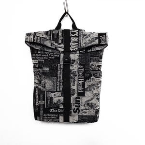 Puede incluir: Una mochila enrollable con un patrón de estampado de periódico en blanco y negro. La bolsa incluye una correa vertical central negra con cierre de hebilla de plástico, dos asas superiores y fragmentos de texto visibles como The Guardian y The Herald.