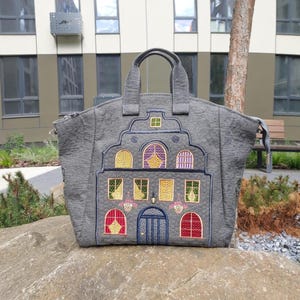 May include: A grey tote bag featuring an embroidered design of a multi-story building with a stepped gable roof. The embroidery includes colorful windows in yellow, red, and purple, a central arched doorway, and two top handles with a zipper closure.