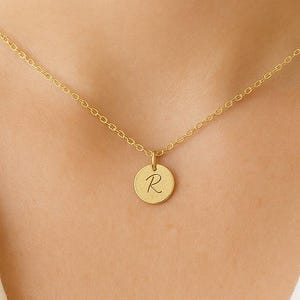 May include: A gold-colored necklace with a delicate chain and a circular pendant. The pendant features the letter "R" in a cursive font. The necklace is displayed against a white background.