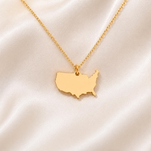 May include: A gold-toned necklace featuring a pendant shaped like the contiguous United States. The pendant is suspended from a delicate, matching gold-toned chain. The surface of the map pendant is smooth and solid, with no internal markings.