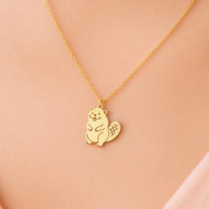 May include: A gold-colored necklace featuring a pendant shaped like a beaver. The beaver pendant has a detailed design, including a cross-hatched tail. The necklace is displayed against a soft pink background.