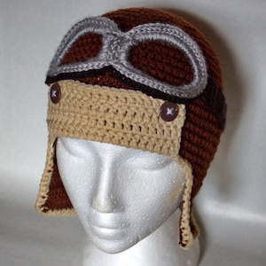 May include: A brown and beige crocheted hat with a gray and brown faux leather aviator goggles design. The hat has two brown buttons on the front and two long brown and beige crocheted straps.