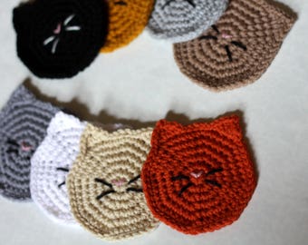 Könnte beinhalten: Ein Set aus sieben gehäkelten Untersetzern in Katzenform in verschiedenen Farben, darunter Grau, Weiß, Beige, Orange und Schwarz. Jeder Untersetzer zeigt ein einfaches Katzen Gesicht mit rosa Nase und schwarzen Schnurrhaaren.