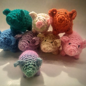 May include: A group of eight crocheted pig toys in various colors, including blue, green, pink, orange, and white. The toys are arranged in a circle, with the blue pig in the front.