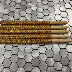 Op de afbeelding: Vier paar eetstokjes met goudkleurige strass-steentjes. De eetstokjes zijn gemaakt van lichtgekleurd hout en zijn versierd met een band van gouden strass-steentjes aan de bovenkant. De eetstokjes zijn gerangschikt op een zilver-grijze zeshoekige tegeloppervlak.