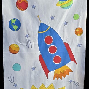 May include: A white fabric with a blue rocket ship, a yellow sun with a smiling face, and colorful planets and stars. The sun has a smiling face.