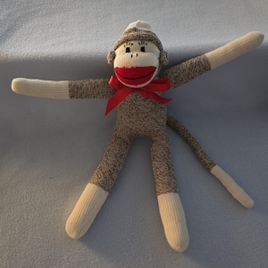 May include: A sock monkey doll crafted from brown and cream-colored knit fabric. The monkey features a bright red stitched mouth, a red ribbon tied around its neck, and long arms and legs with cream-colored ends. It has a long tail and a hat.