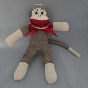 May include: A brown and white sock monkey with a red bow tie. The monkey has a white face with black eyes and a red mouth.