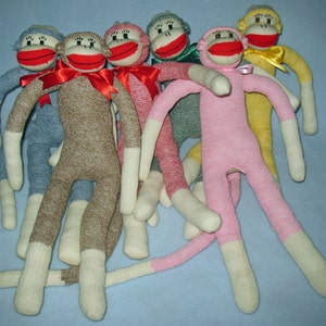 May include: Six sock monkeys in various colors, including blue, brown, pink, red, yellow, and green. The monkeys are made from knitted socks and have embroidered faces with red lips. They are arranged in a group on a light blue surface.