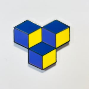 May include: A blue and yellow geometric pin with a three-dimensional design. The pin is made of metal and has a shiny finish.