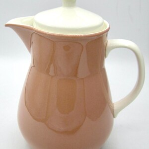 May include: A ceramic jug with a lid, handle, and spout. The jug is a light brown colour with a white lid, handle, and rim. The jug has a glossy finish and is likely used for serving drinks.