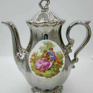 May include: Silver teapot with a white porcelain design of a couple in a garden setting.