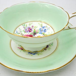May include: A vintage teacup and saucer set. The cup and saucer are a pale green with gold edging. The interior of the cup and the centre of the saucer feature a floral design with purple, blue, and pink flowers.