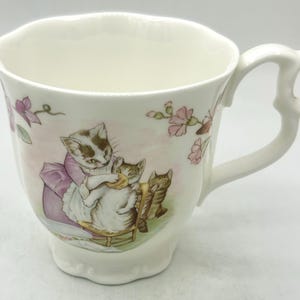 May include: A cream-colored porcelain teacup with a scalloped rim and an ornate handle. The exterior features a colorful illustration of three cats, with one cat wearing a purple dress, alongside pink floral accents and green leaves.